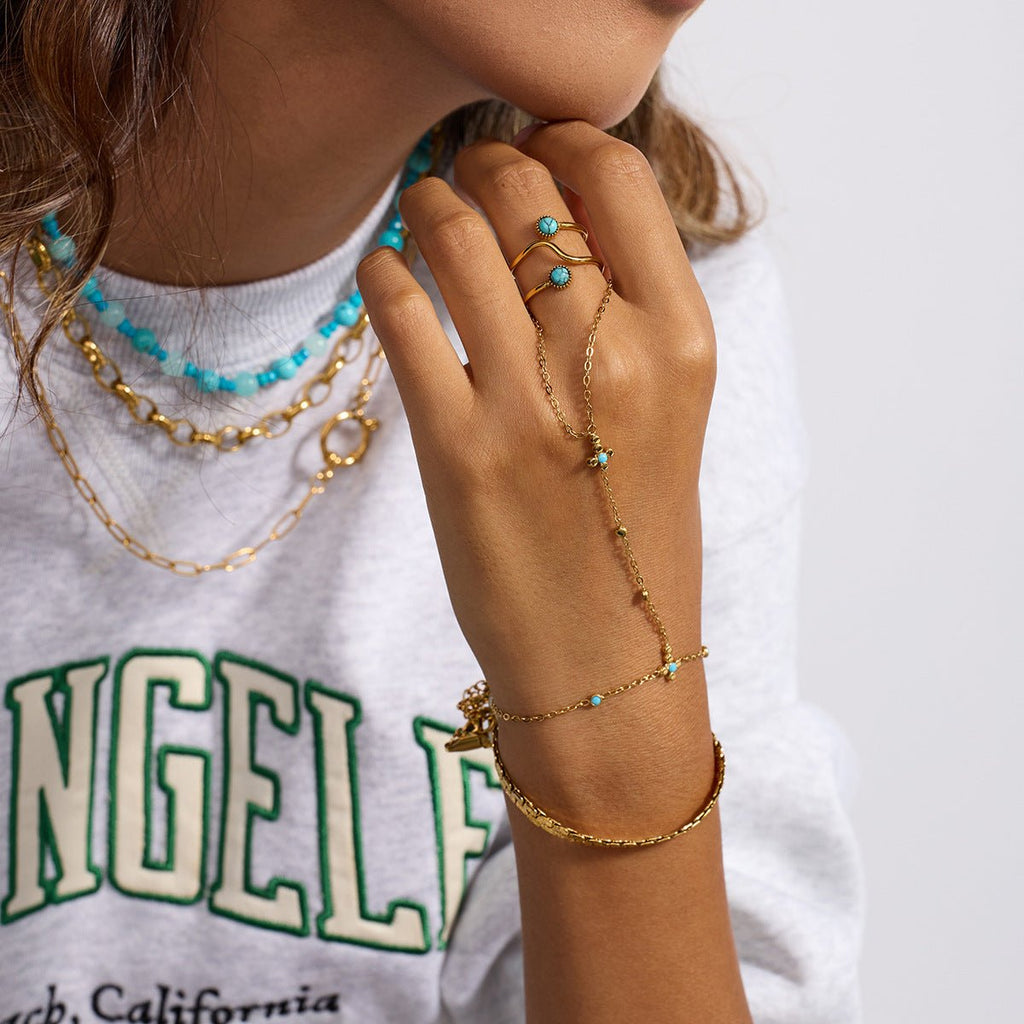 Chaîne de doigt en acier inoxydable doré avec perles turquoise et petite croix, portée avec bracelet doré assorti.