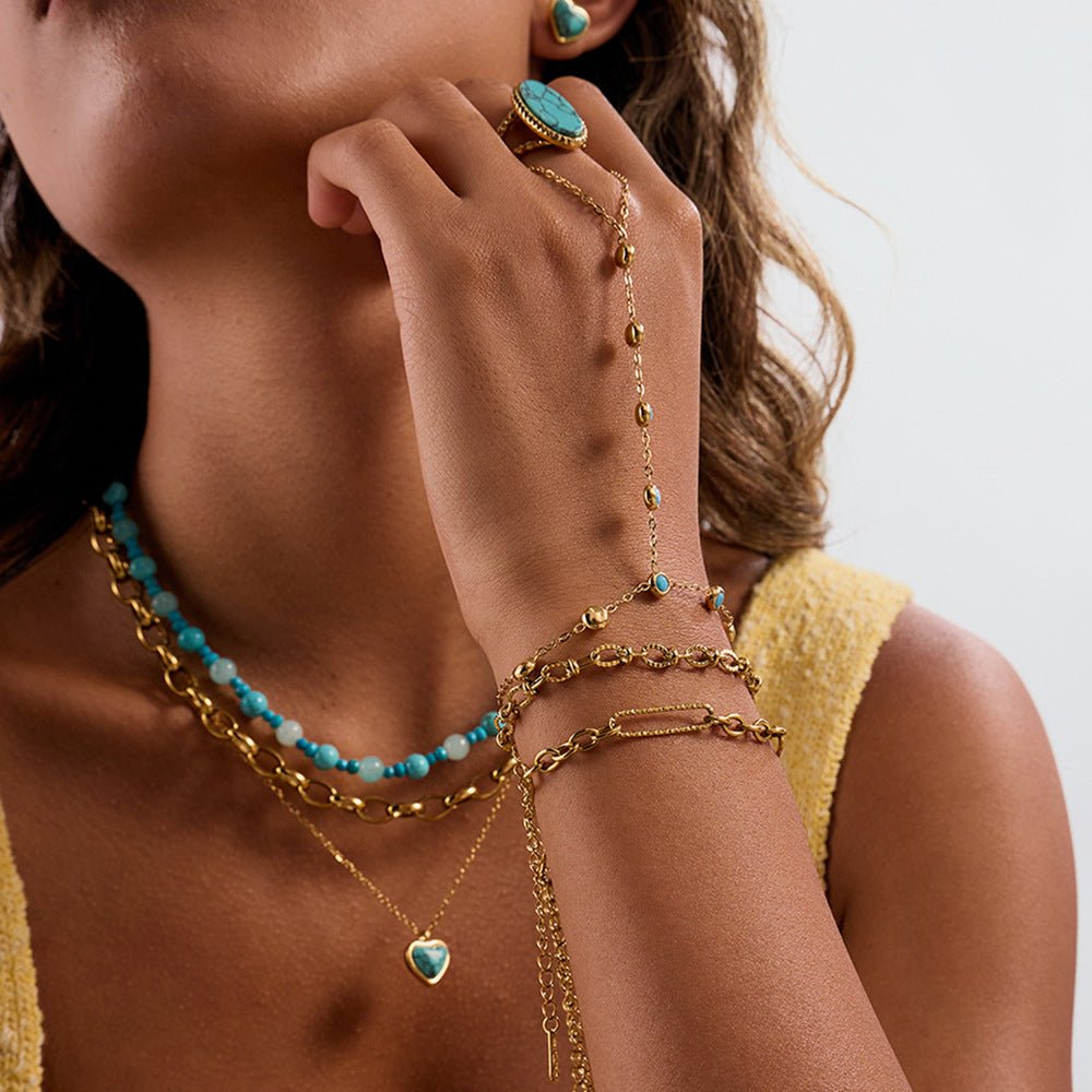 Chaînette de main dorée en acier inoxydable avec pierres turquoise ovales, style Sandy, portée avec bracelets et collier.