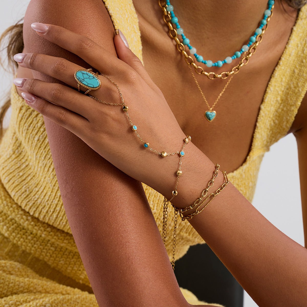 Chaînette de main dorée en acier inoxydable avec petites pierres turquoise et bague assortie sur la main féminine.