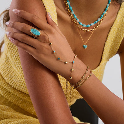 Chaînette de main dorée en acier inoxydable avec petites pierres turquoise et bague assortie sur la main féminine.