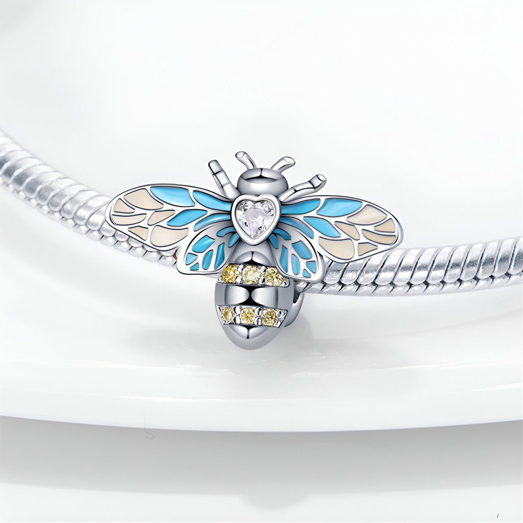 Charm abeille argentée avec ailes bleues et cœur en cristal transparent, détails jaunes sur le corps.