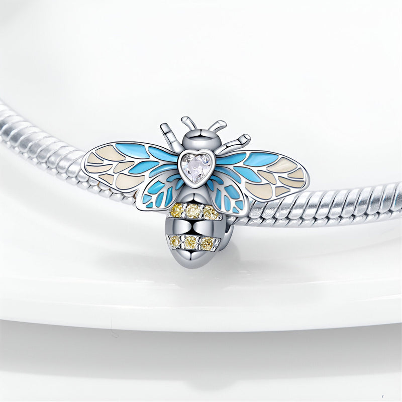 Charm abeille argentée avec ailes bleues et cœur en cristal transparent, détails jaunes sur le corps.