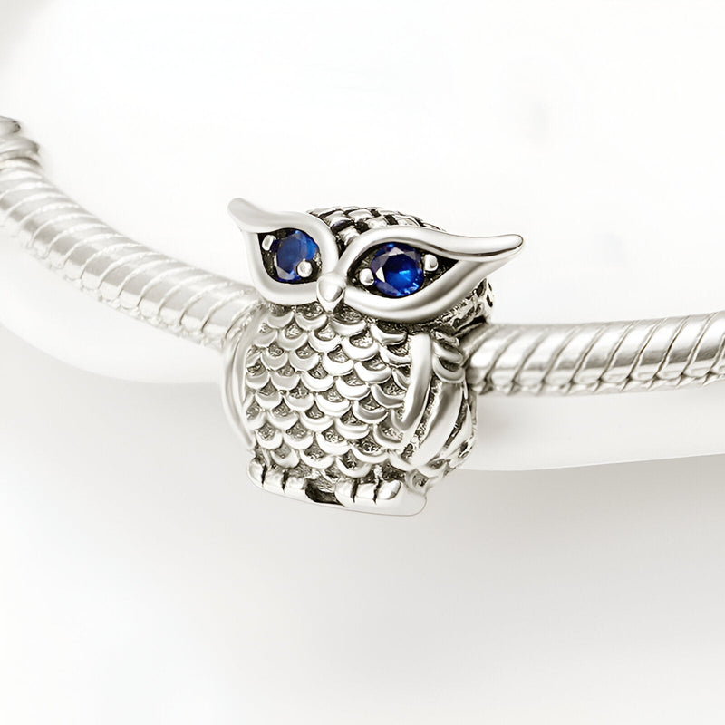 Charm hibou en argent avec yeux bleus en cristal, détails de plumes texturées, style élégant.