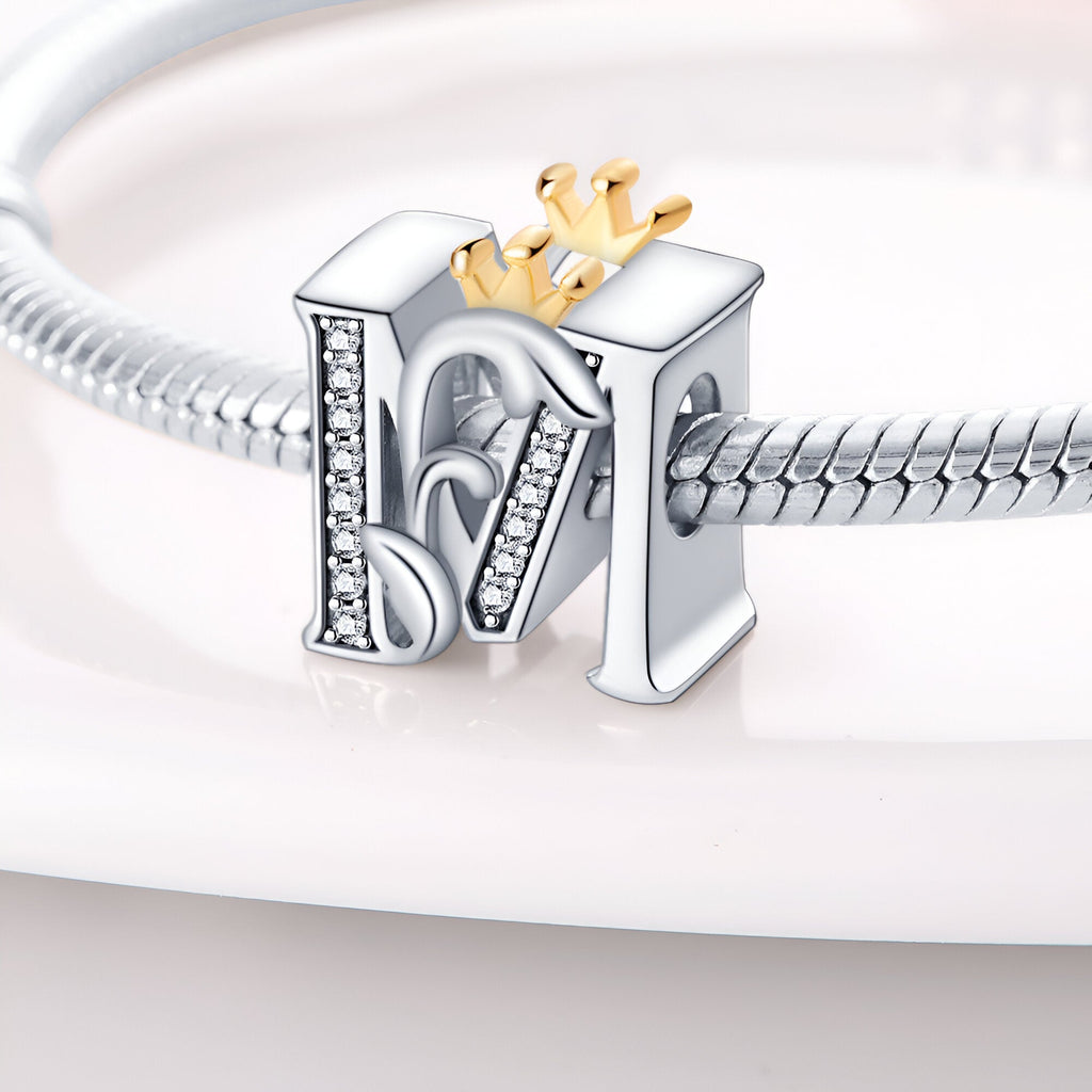 Charm lettre M en argent avec détails en strass et petite couronne dorée sur bracelet serpent en métal.