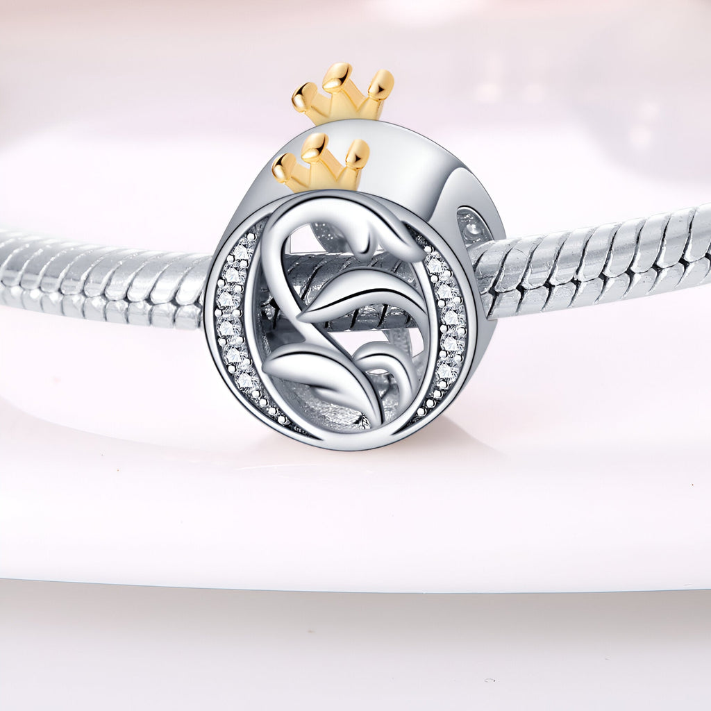 Charm lettre O argenté avec couronne dorée et détails feuille, serti de petits cristaux blancs.
