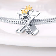 Charm en forme de lettre X argentée avec couronne dorée et détails en cristaux sur bracelet serpent argenté.
