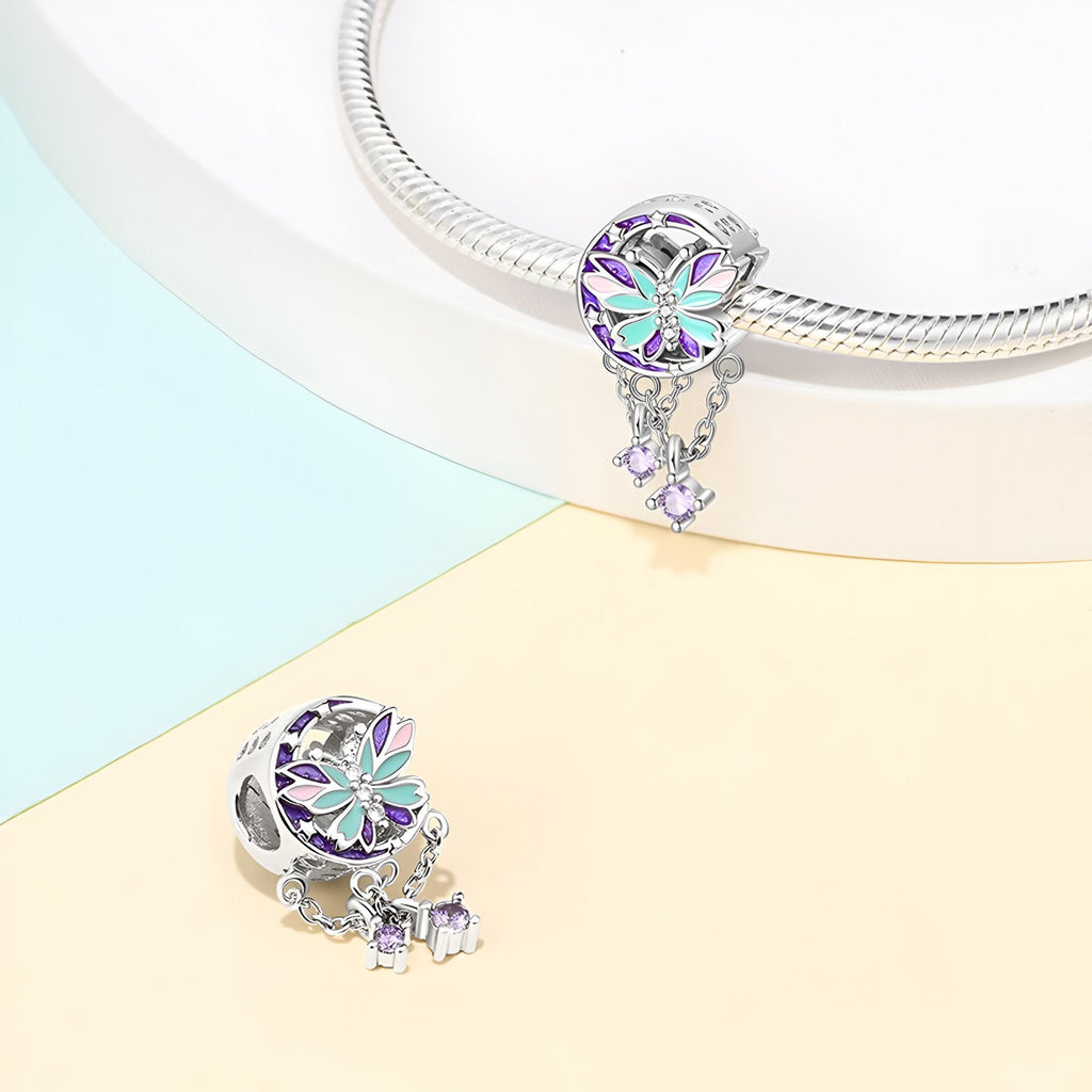 Charm argenté en forme de papillon avec détails colorés violets et bleus, orné de petites pierres pendantes.