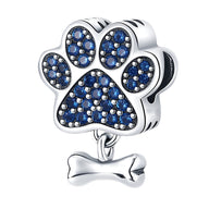 Charm argent en forme de patte de chien ornée de petits cristaux bleus avec os suspendu.