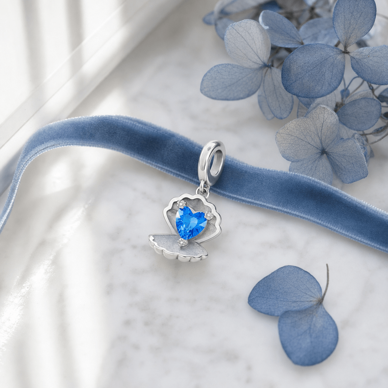 Charm Conchilia coquillage cœur bleu posé sur marbre blanc avec ruban velours bleu et pétales d'hortensia