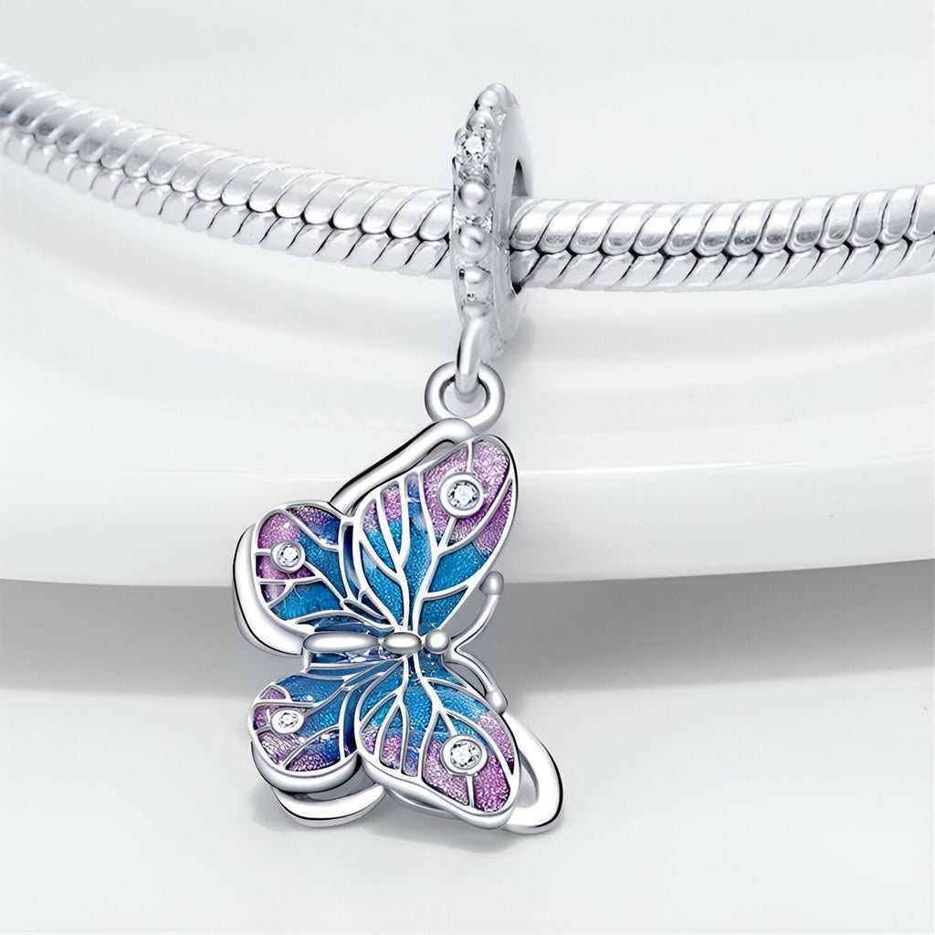 Pendentif papillon bleu et rose en argent avec détails nacrés et pierres scintillantes attaché à un bracelet serpent argenté.