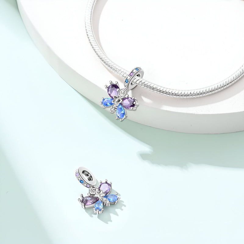 Pendentif papillon en métal argenté avec cristaux bleu et violet attaché à un bracelet serpent blanc.