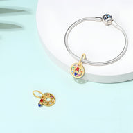 Charm pendentif pinceau doré avec détails colorés et strass bleus sur bracelet argenté fin.