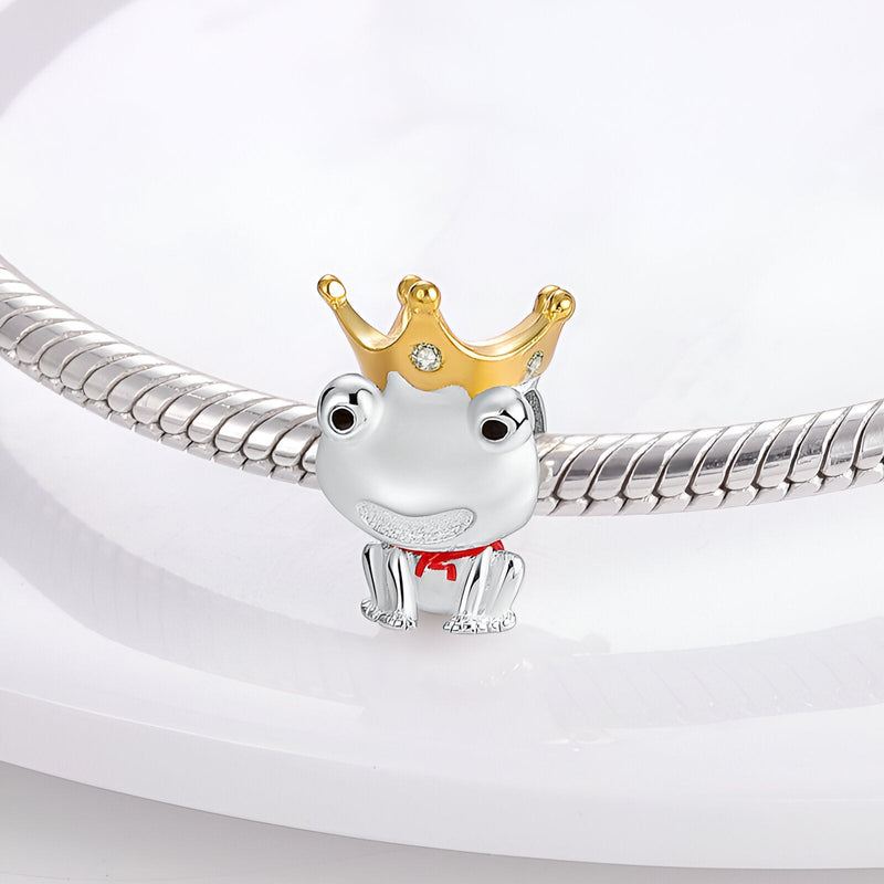Charm grenouille argentée avec couronne dorée et nœud rouge, yeux noirs brillants.
