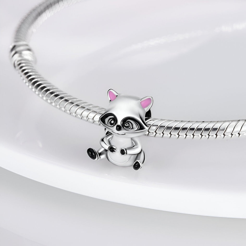 Charm raton laveur argenté avec détails noirs et oreilles roses, suspendu à un bracelet serpent argenté.