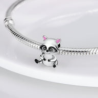 Charm raton laveur argenté avec détails noirs et oreilles roses, suspendu à un bracelet serpent argenté.