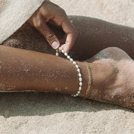 Duo de bracelets de cheville or, perles d'eau douce et chaîne fine en acier inoxydable sur peau bronzée avec sable.