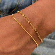 Duo de bracelets fins en acier inoxydable doré, lisse et chaîne ovale, porté au poignet.