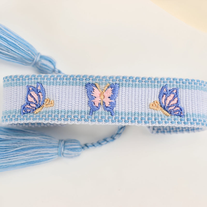 Bracelet en coton tissé bleu clair avec motif papillons rose et bleu, finition pompons.