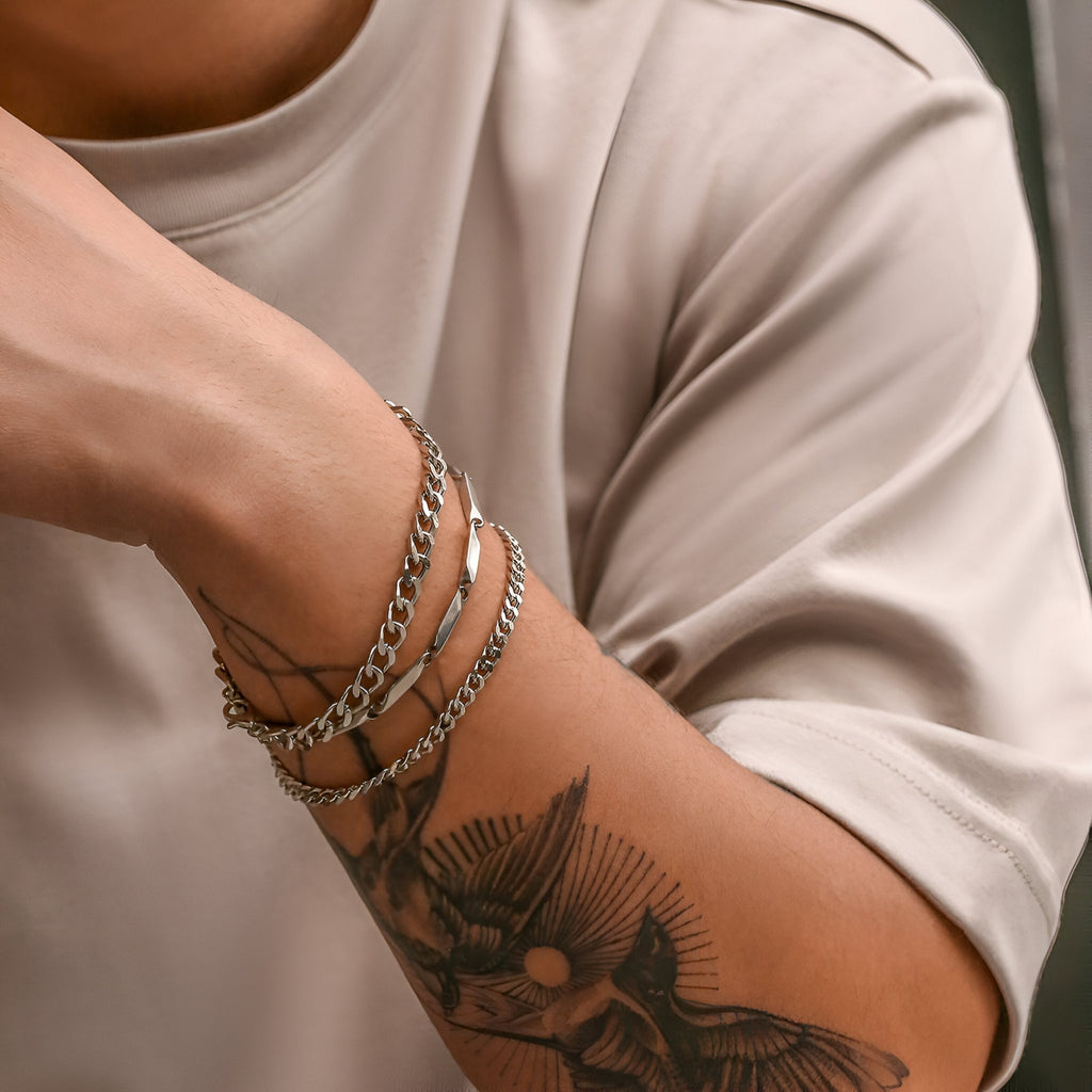 Trio de bracelets maille en titane argentés portés sur poignet masculin avec tatouage noir detailed.
