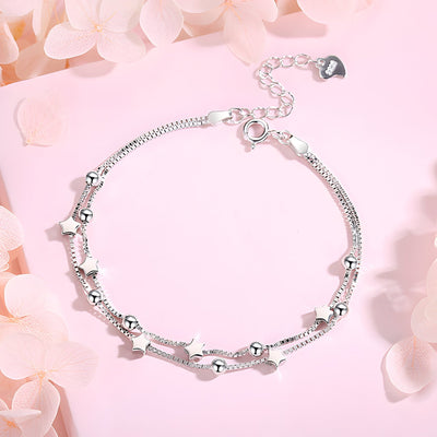 Bracelet fin en argent sterling avec petits charms étoiles et chaînes délicates sur fond rose pâle.