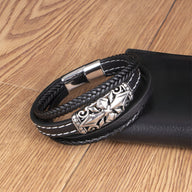 Bracelet multi-rangs noir en cuir tressé avec motif croix argentée et fermoir magnétique métallique.