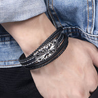 Bracelet multi-rangs noir en cuir avec motif croix argentée détaillée, coutures blanches visibles.