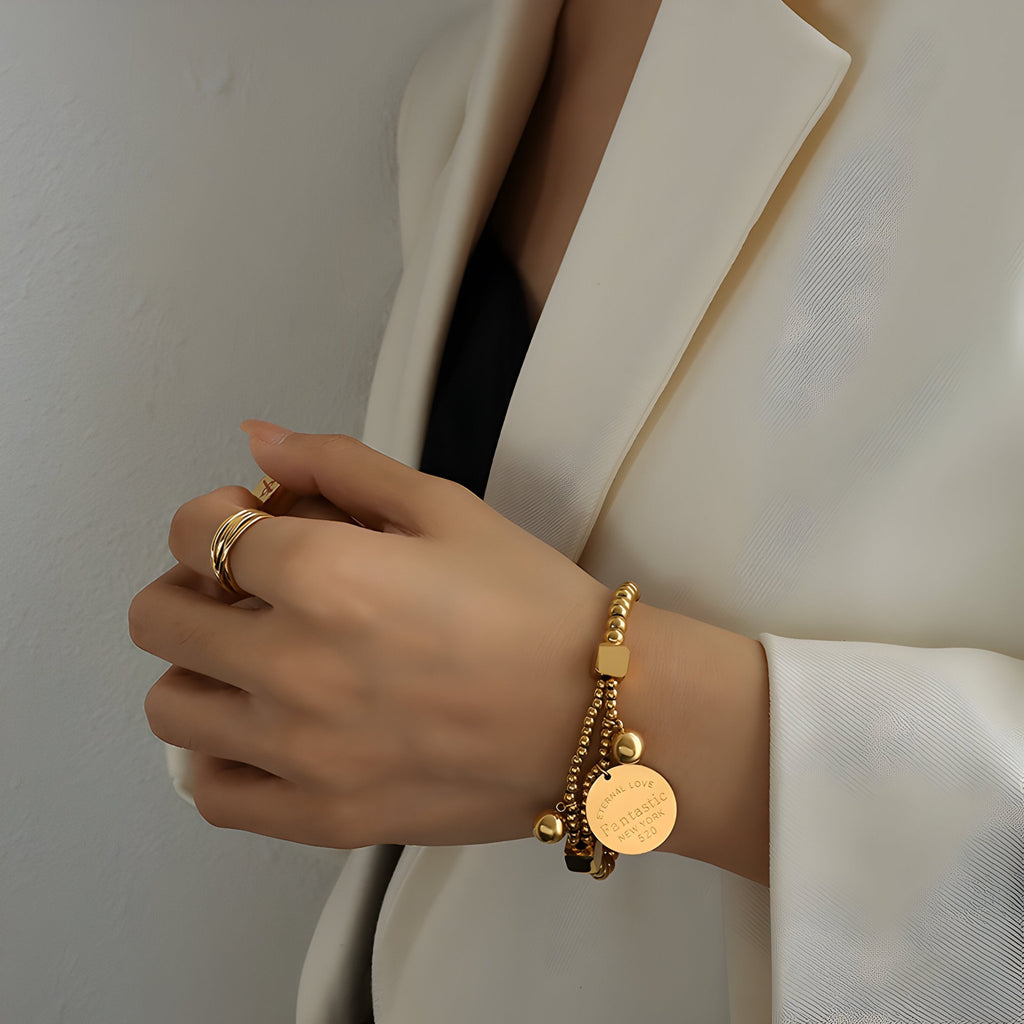 Bracelet en titane doré avec perles rondes et médaillon gravé, porté au poignet féminin.