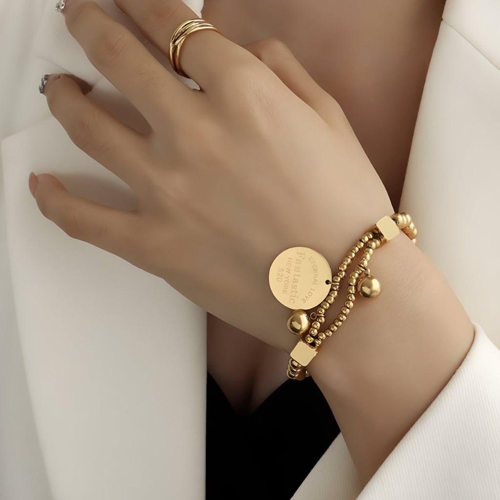 Bracelet en titane plaqué or avec perles dorées et médaillon rond gravé, style Melvina, porté au poignet féminin.