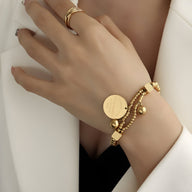 Bracelet en titane plaqué or avec perles dorées et médaillon rond gravé, style Melvina, porté au poignet féminin.