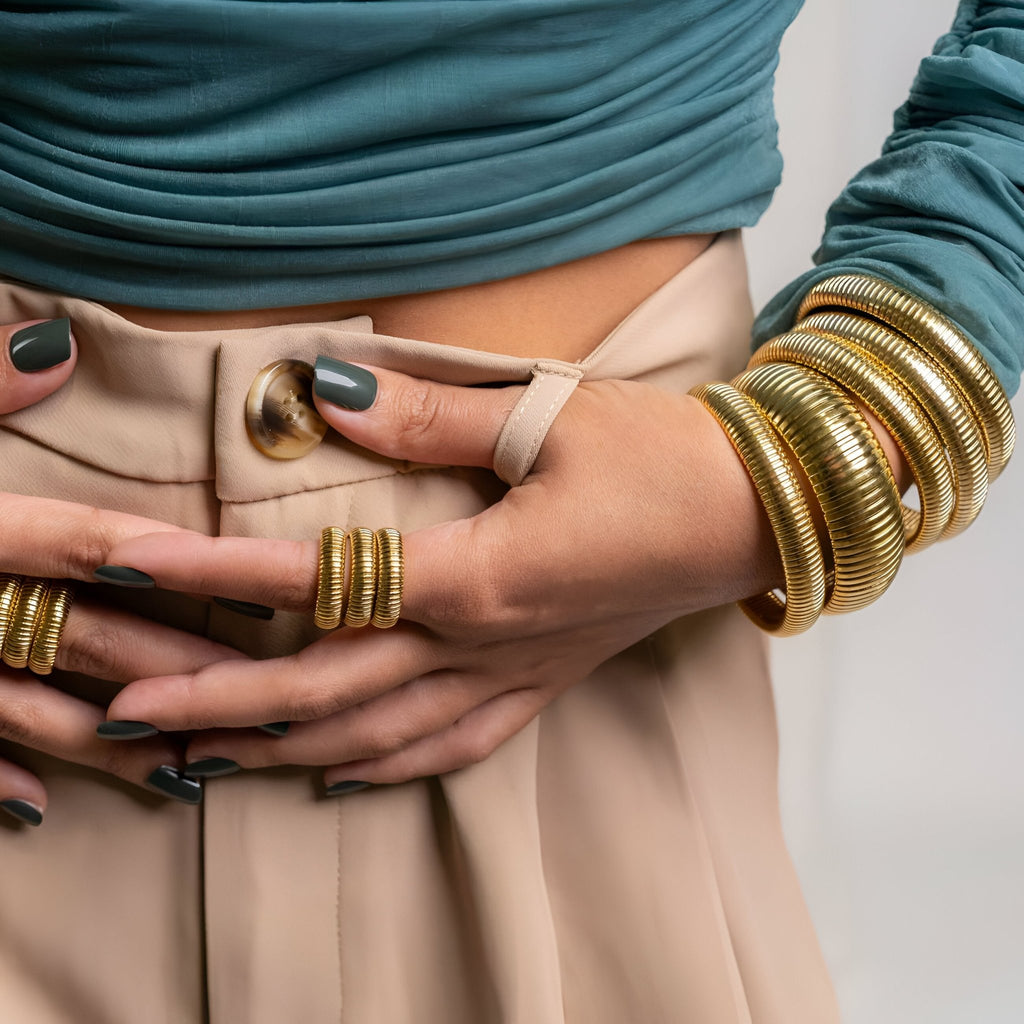 Bracelet souple en titane plaqué or modèle Mossie avec multiples joncs texturés porté avec bague assortie et pantalon beige.