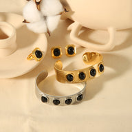 Deux bracelets joncs en or et argent avec pierres naturelles noires et deux bagues assorties sur fond beige.