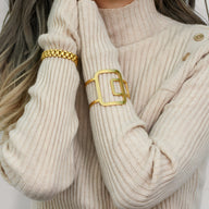 Bracelet manchette doré avec design géométrique carré porté sur pull côtelé beige.