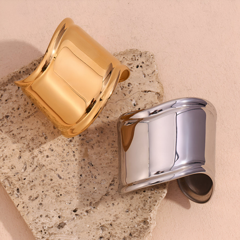 Bracelet manchette large en acier inoxydable finition or brillant et argenté posé sur pierre beige texturée.