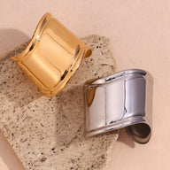 Bracelet manchette large en acier inoxydable finition or brillant et argenté posé sur pierre beige texturée.