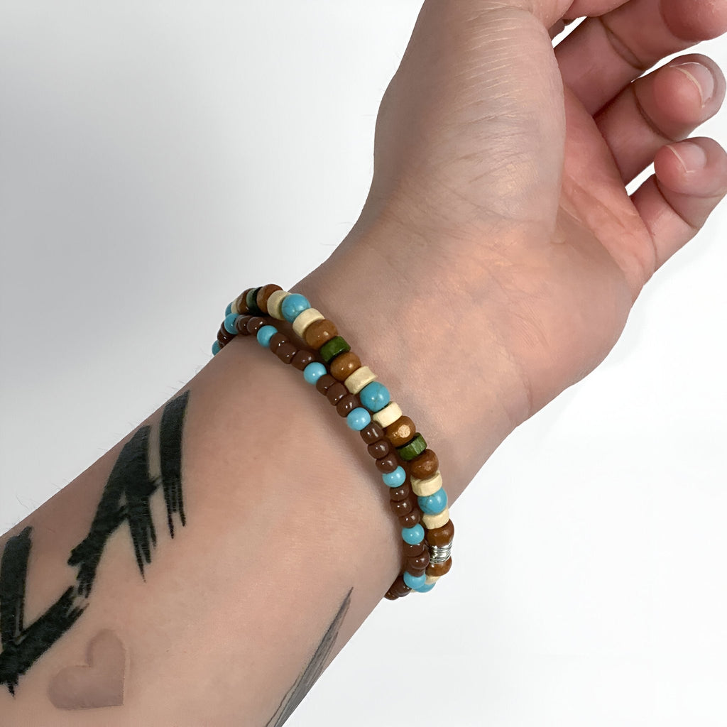 Ensemble de deux bracelets homme en perles de bois marron, turquoise et blanches sur poignet tatoué.